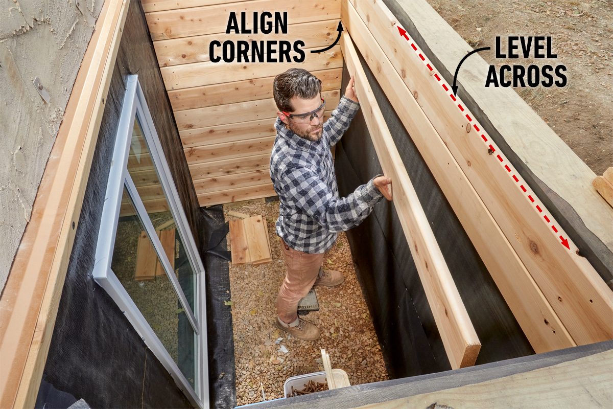 A person positions wooden beams while ensuring they are aligned and leveled inside a gravel-floored construction space, surrounded by wooden walls and a window.