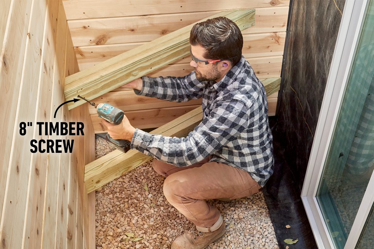 A man kneels, using a power drill to secure wooden beams together in a gravel area between wooden walls and a window.