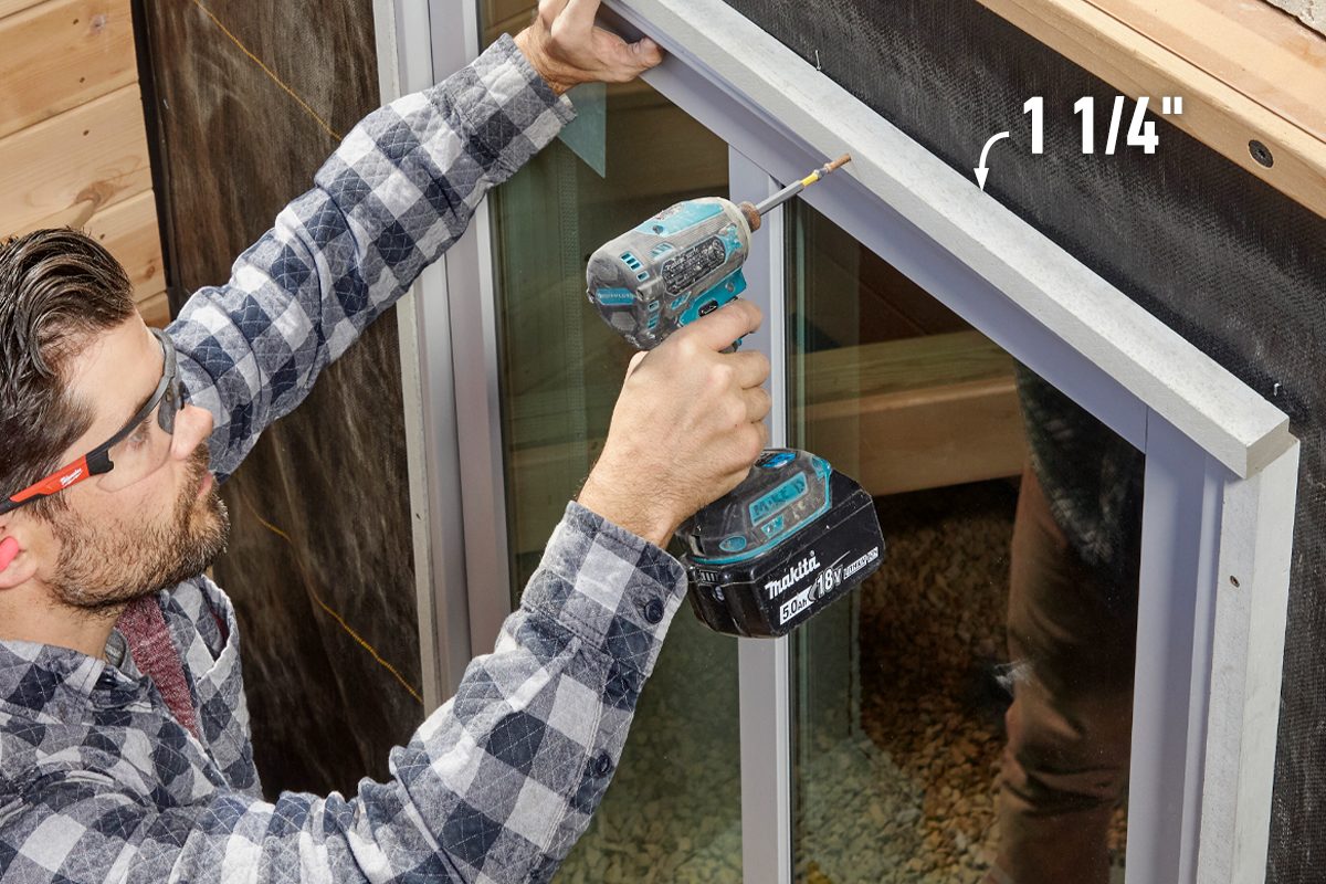A man uses a power drill to install a frame above a window, surrounded by wooden structures and gravel on the ground.