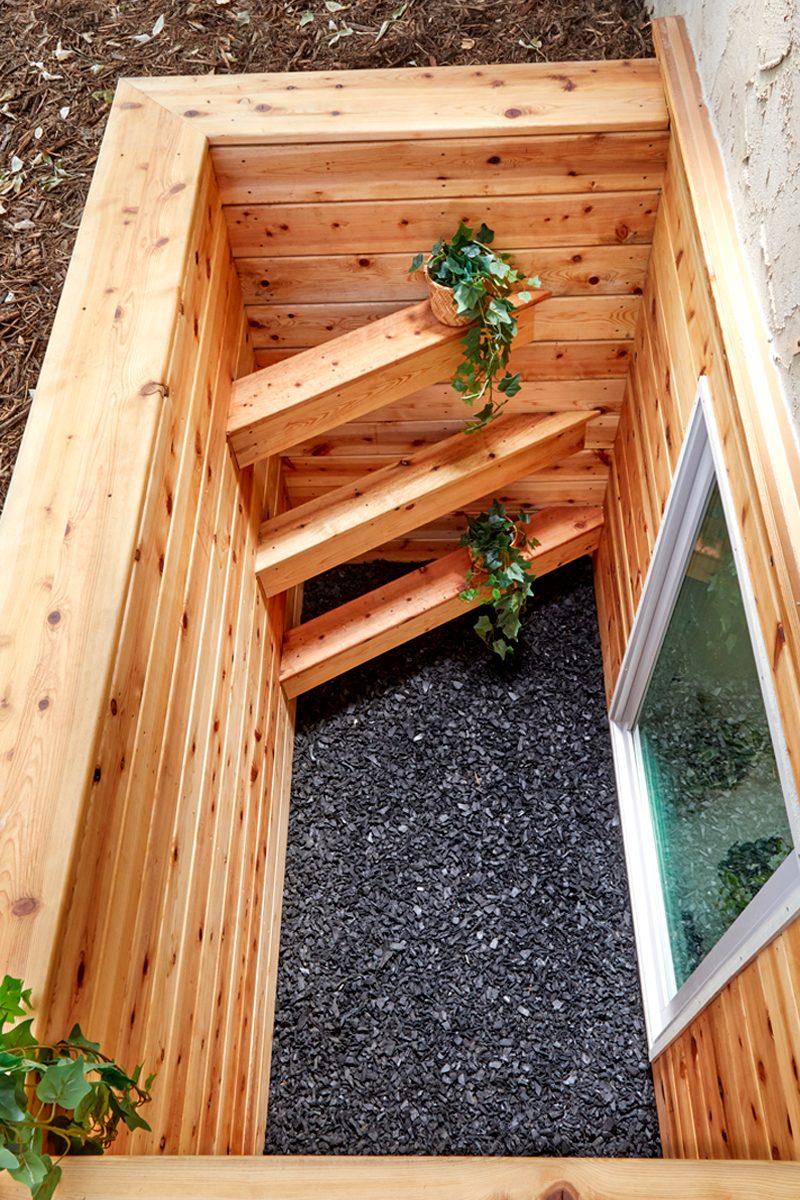 A wooden stairwell features steep steps, with potted plants and a window overlooking a gravel-filled space below, surrounded by earthy materials.