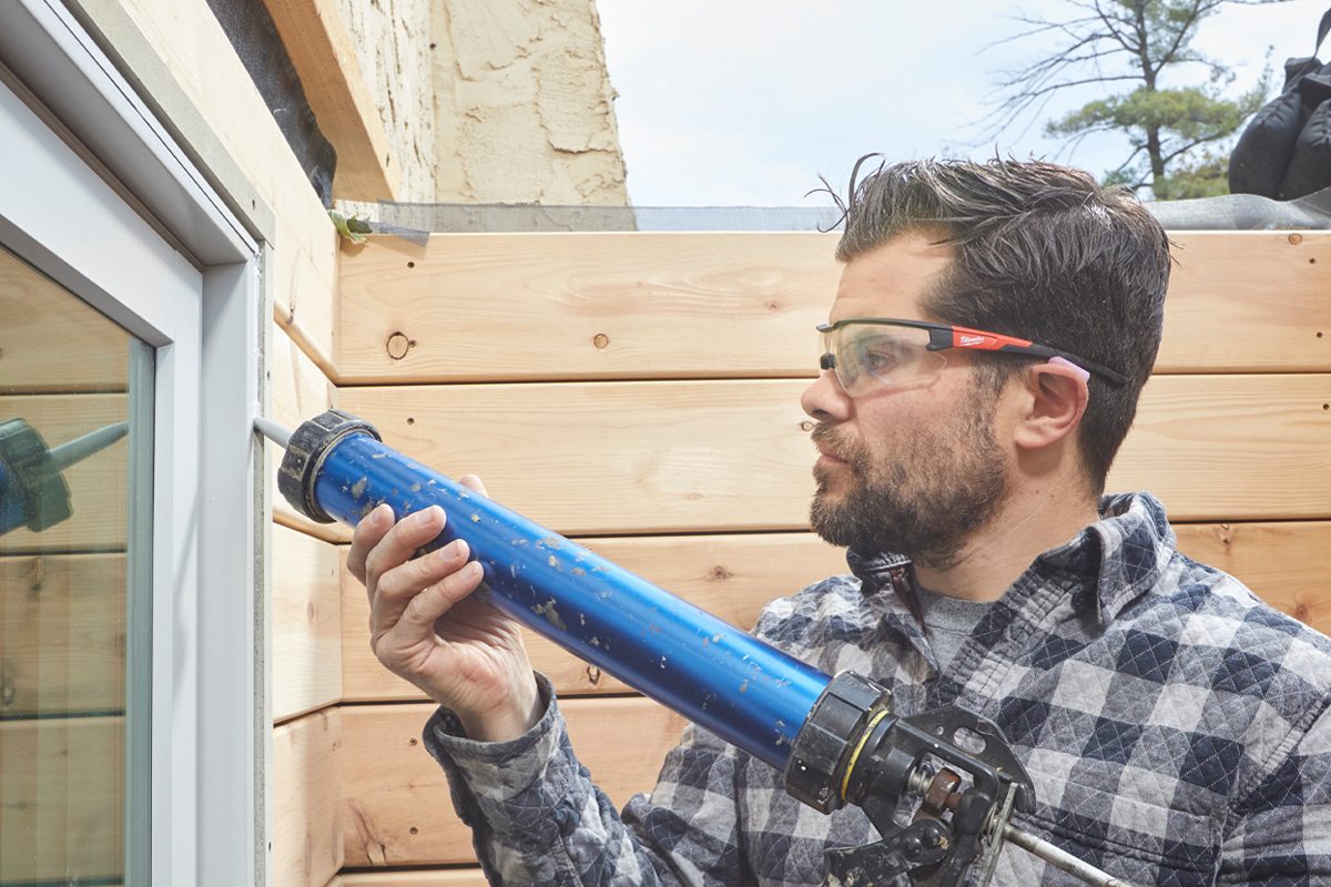 A man applies caulk using a large blue caulking gun to seal gaps around a window frame, surrounded by wooden siding and a partially finished building.