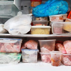 Multiple containers filled with frozen food items are haphazardly stacked inside a freezer, creating a cluttered and disorganized appearance.