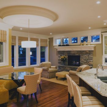 A cozy kitchen-living area features a glass table, soft upholstered seating, a stone fireplace, and large windows, illuminated by pendant lighting.