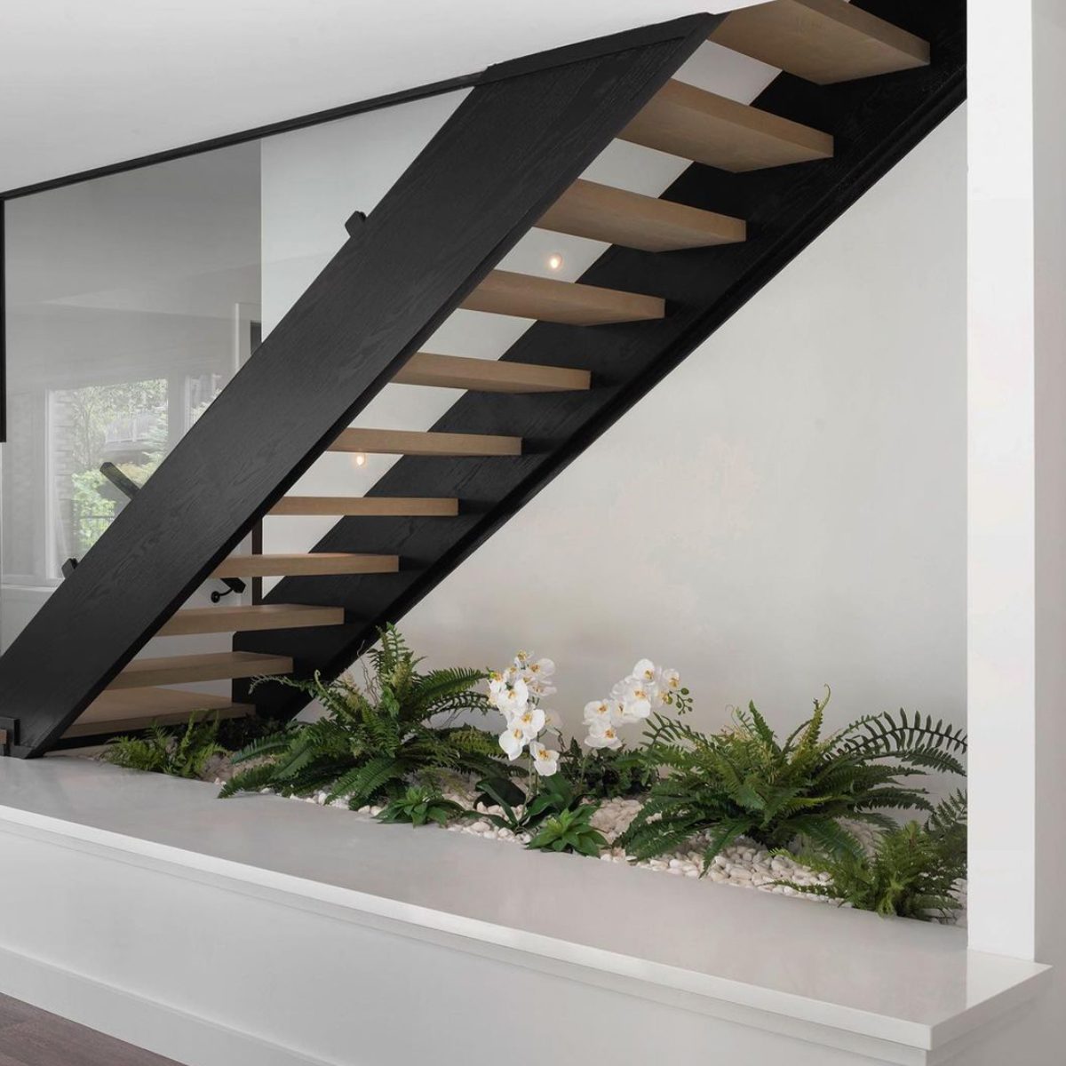 A modern staircase with black railings ascends above a decorative plant arrangement featuring white orchids and ferns, set against a minimalist wall.