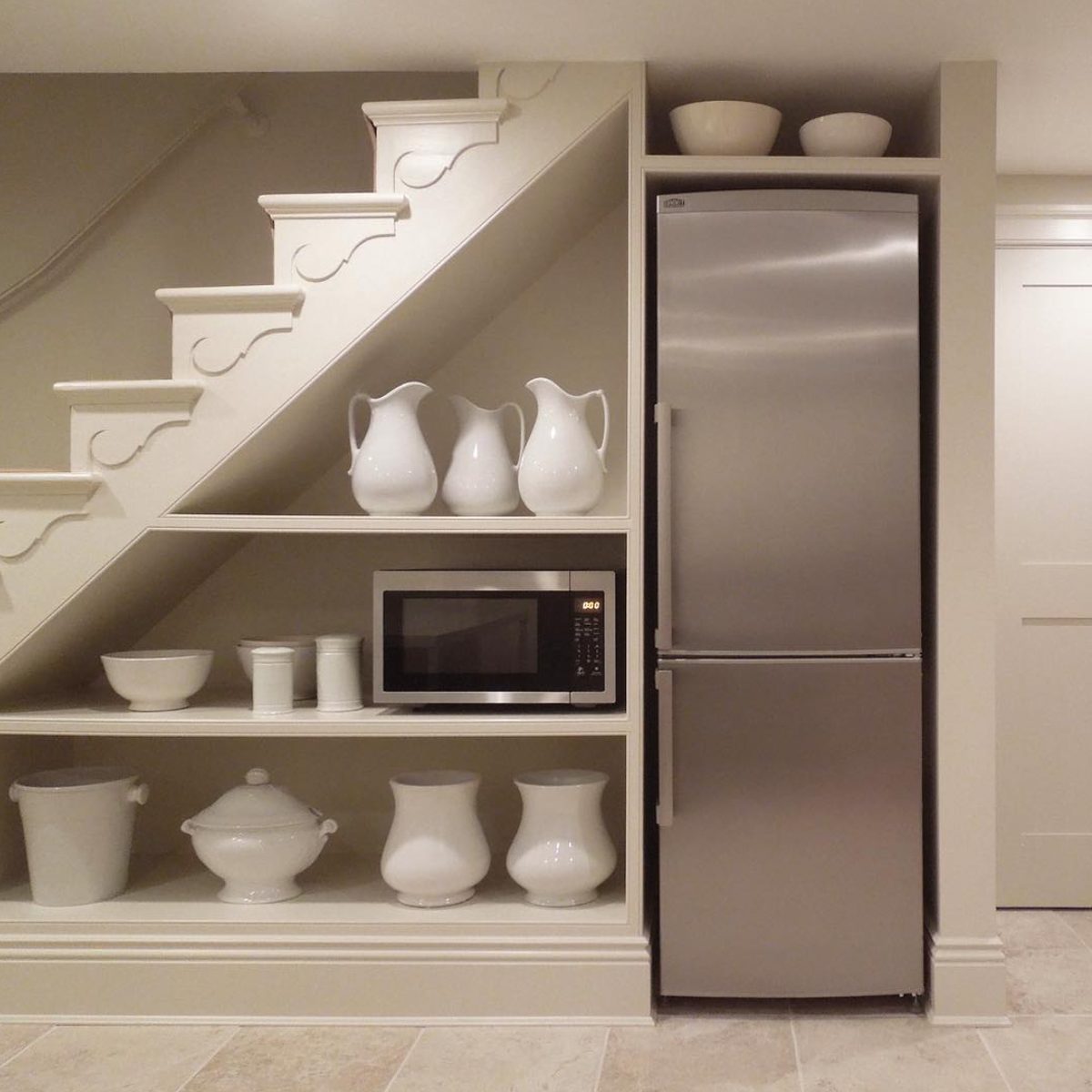Shelving unit beside a stainless steel refrigerator holds white ceramic pitchers, bowls, and a microwave, situated under a staircase in a neutral-toned room.