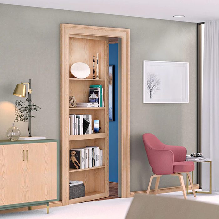 A wooden bookshelf displays various books and decor while a pink chair sits beside a side table in a modern, softly lit room.