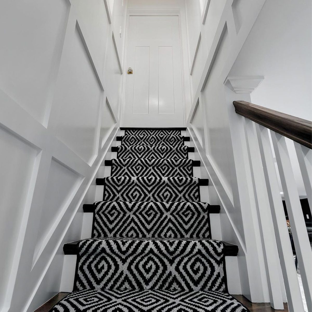 A black and white patterned carpet covers stairs ascending to a closed door, framed by white walls and a wooden railing on the right.