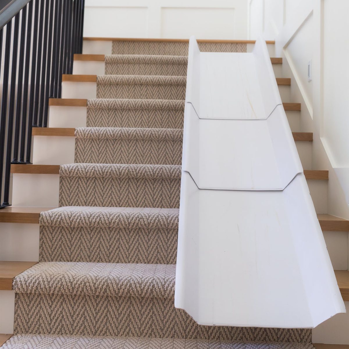 A white slide extends down a carpeted staircase, offering a playful descent. The stairs have wooden edges and a textured pattern on the carpet.