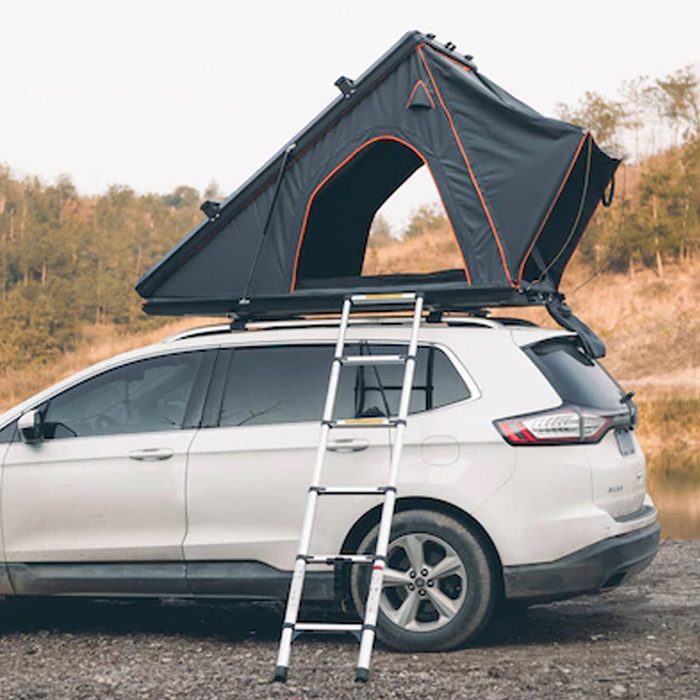 A rooftop tent is opened atop a white vehicle, with a ladder leaning against it, set in a natural landscape with trees in the background.