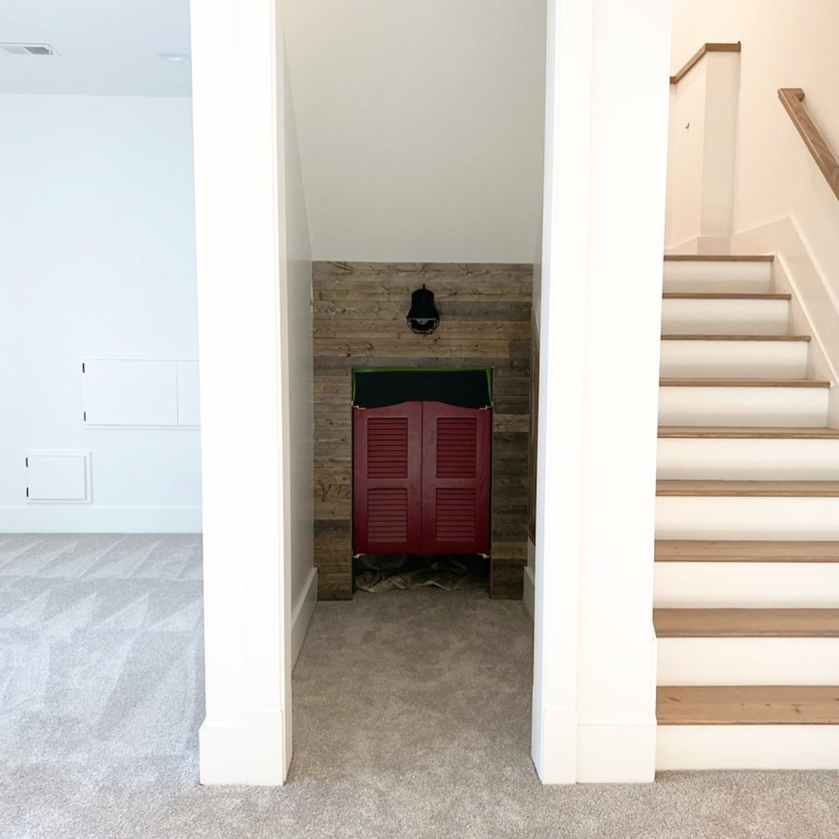A red shuttered door stands at the end of a carpeted hallway, with wooden paneling and a light fixture above, flanked by a staircase.