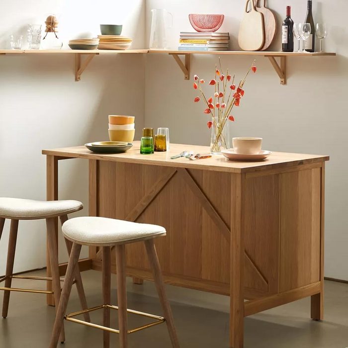 A wooden table holds various dishes and glasses, while two upholstered stools sit nearby; light filters in, creating a cozy, inviting atmosphere.