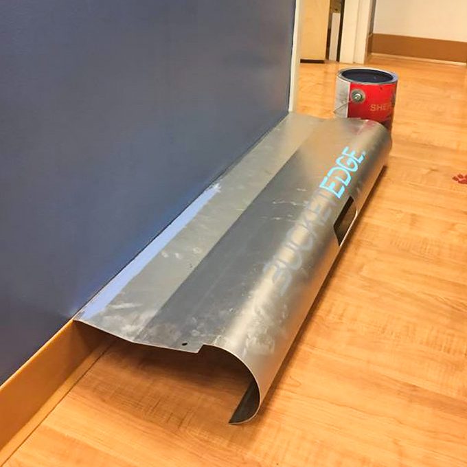 A metal edge trim rests against a blue wall, partially covering a wooden floor, beside a can of paint.