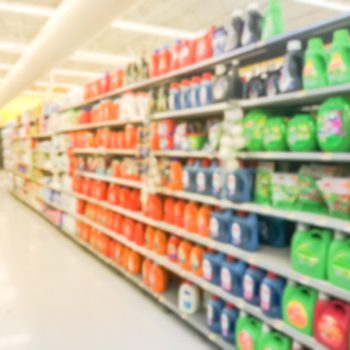 Rows of colorful liquid detergent containers are neatly arranged on shelves, with a bright store ambiance in the background, suggesting a retail environment.