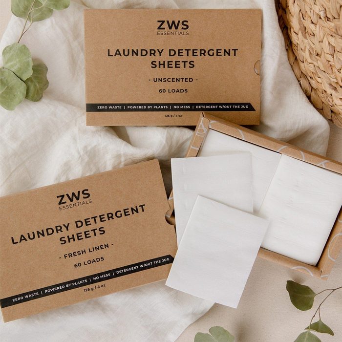 Two boxes of unscented laundry detergent sheets sit on a textured cloth with a few sheets partially visible, surrounded by greenery and a woven basket.