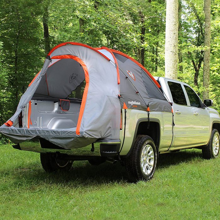 A truck with a tent attached to its bed is parked in a grassy area surrounded by trees, suggesting a camping or outdoor setting.