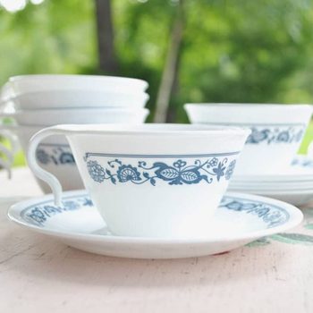 A white tea cup and saucer, adorned with blue floral designs, sit on a wooden table, surrounded by stacked cups, against a blurred green backdrop.