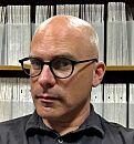 A person with short, light-colored hair and glasses gazes thoughtfully, positioned in front of a background filled with neatly arranged, stacked documents.
