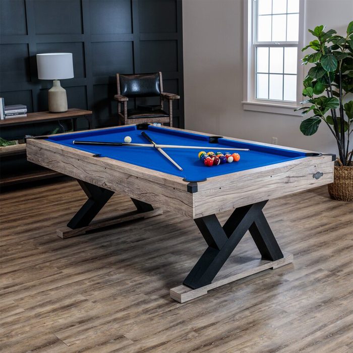 A billiards table with a blue felt surface features a cue ball and numerous colored balls arranged for play, set in a stylish room with wooden decor.