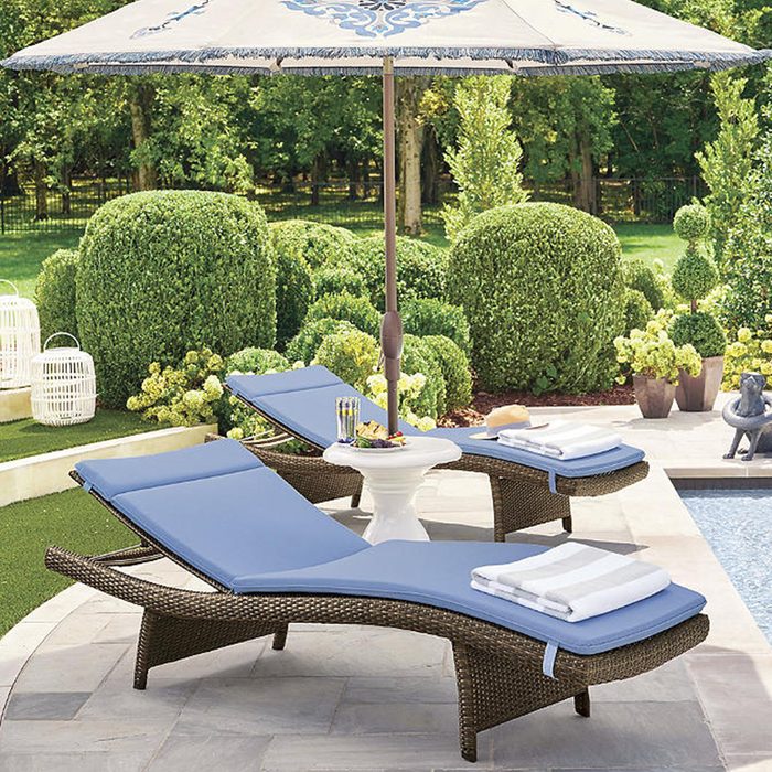 Two lounge chairs with blue cushions are positioned by a pool, shaded by an umbrella, surrounded by neatly trimmed hedges and decorative plants.