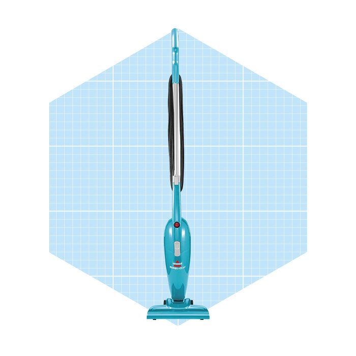 A turquoise vacuum cleaner stands upright against a light blue grid background, ready for cleaning surfaces. Its design features a prominent handle and a red power button.