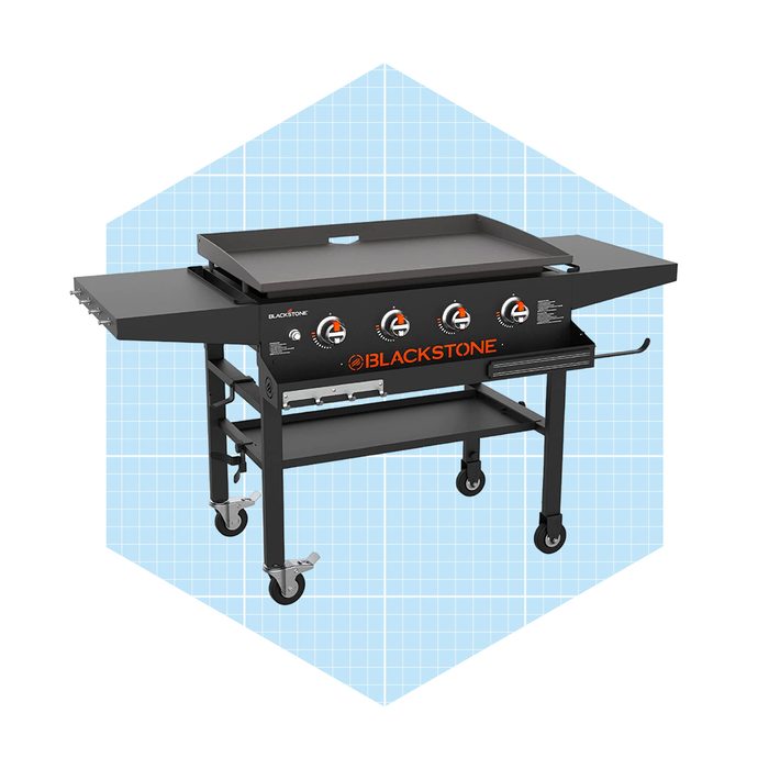 A large, black outdoor griddle with four knobs sits on a wheeled frame, positioned against a light blue geometric background.