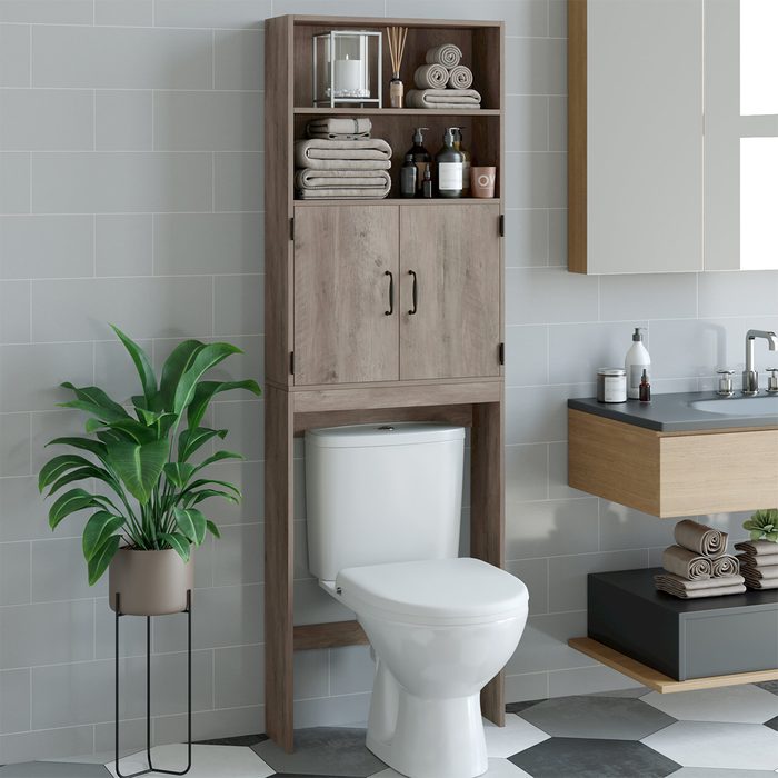A wooden cabinet rests above a white toilet, storing towels and toiletries in a modern bathroom with gray tiles and a green potted plant nearby.