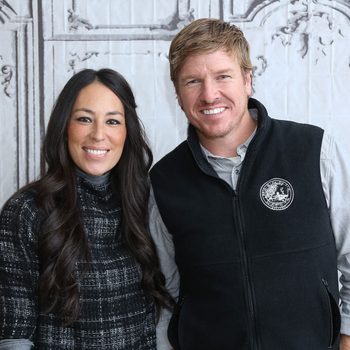 Two smiling individuals stand closely together, dressed casually, in front of a decorative backdrop that suggests a vintage setting, with intricate designs.