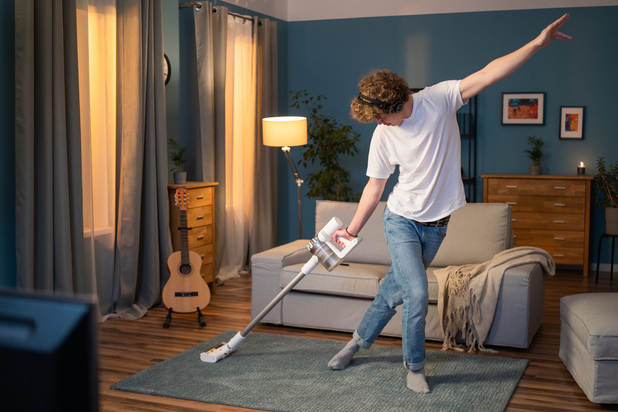 A boy is cleaning the living room in the evening. The man has fun vacuuming the carpet in the room, listening to music on wireless headphones and dancing with the vacuum cleaner
