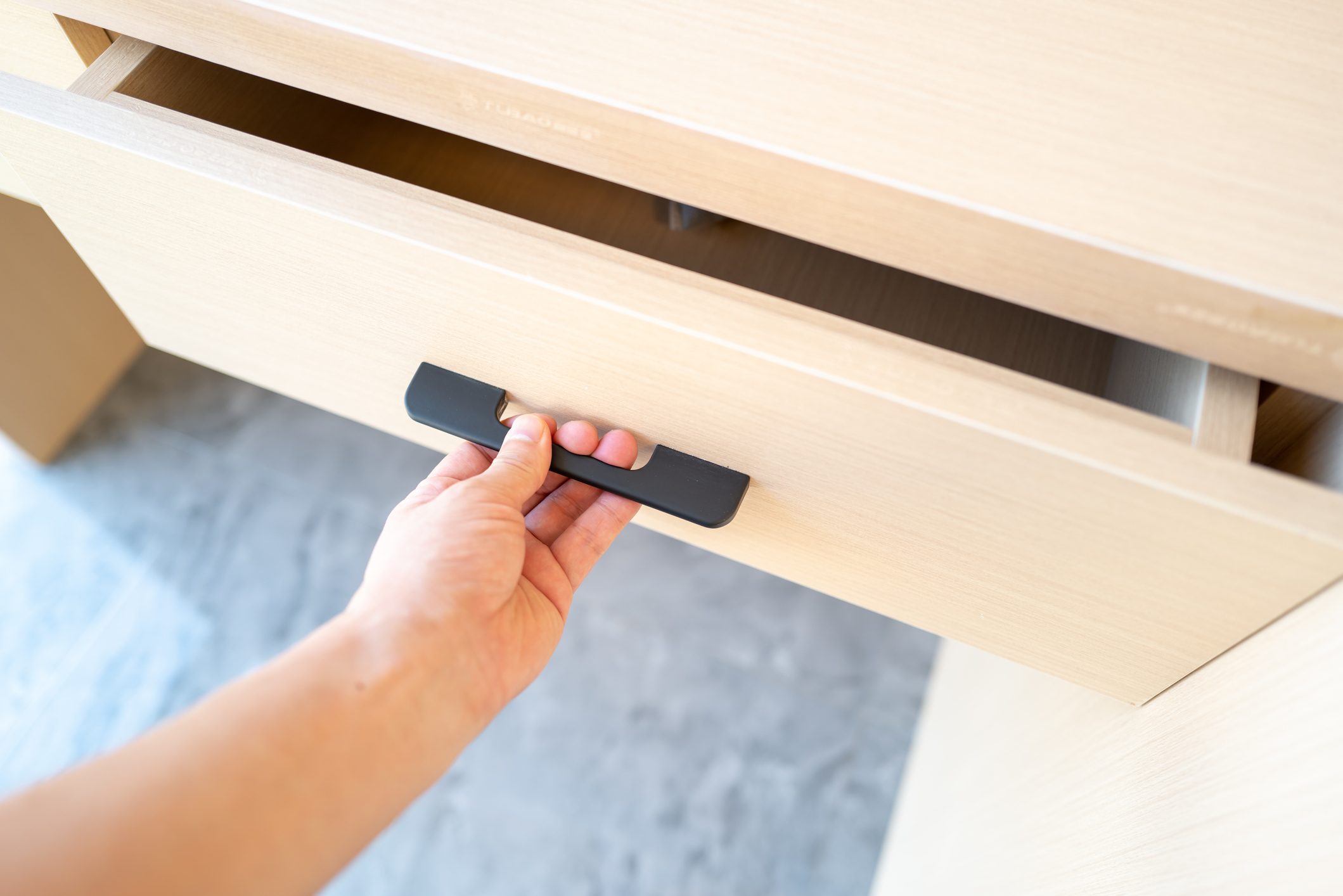A hand grips a black drawer handle, pulling it open on a light wood cabinet, revealing a partially opened drawer against a smooth, grey surface.