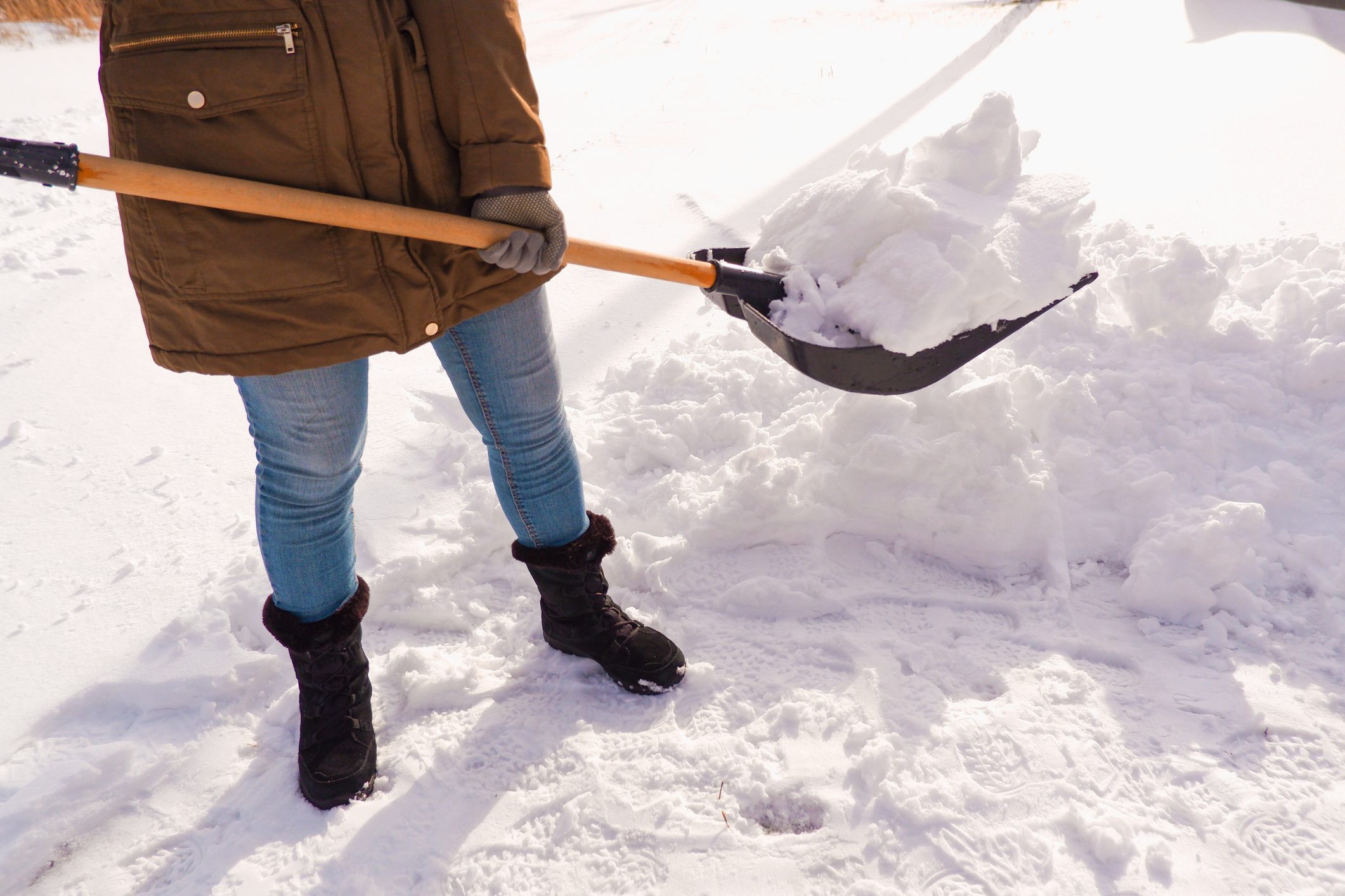 A person shovels snow with a black shovel, standing on a snowy ground, wearing a brown coat, blue jeans, and black boots.