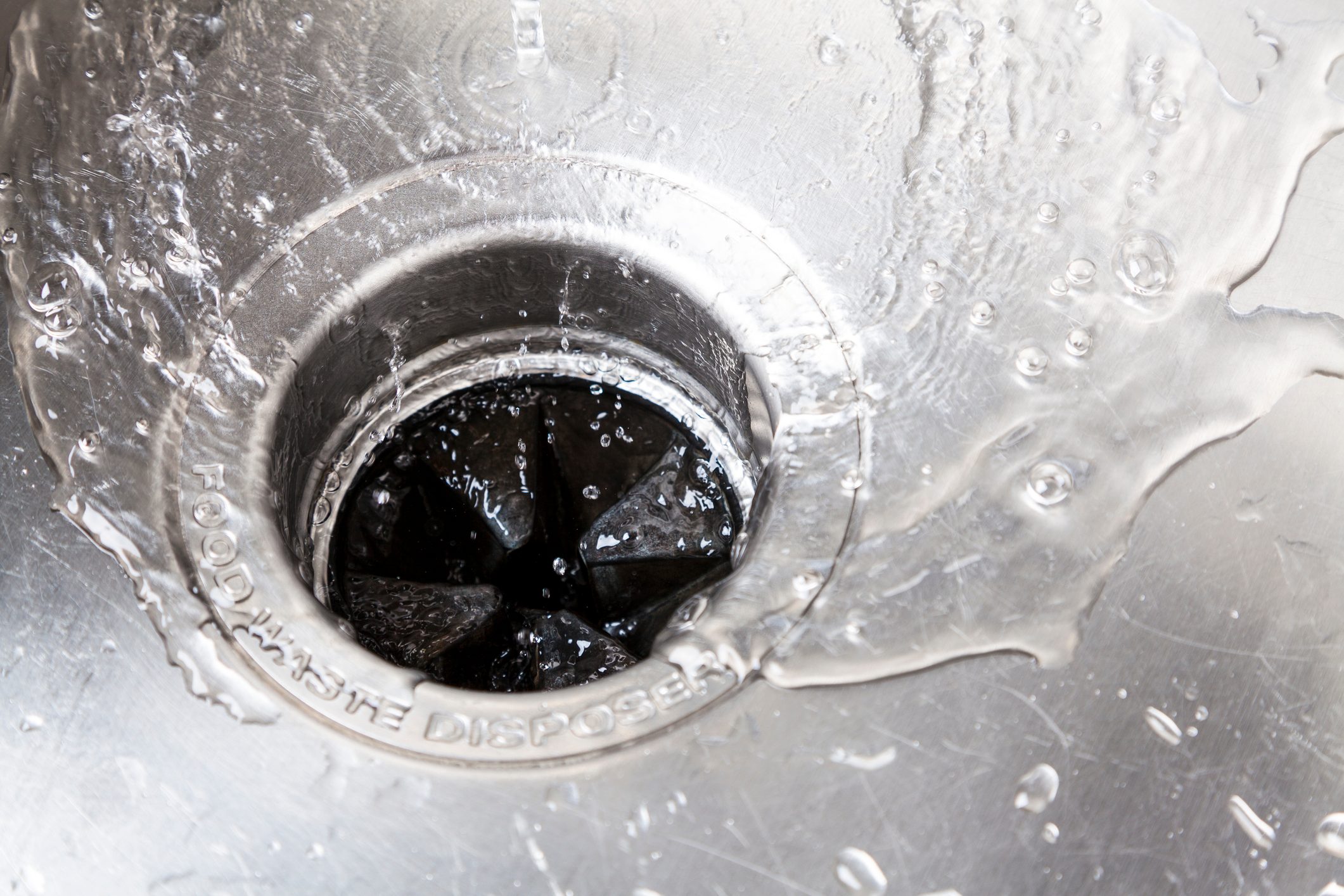 A stainless steel sink drains water, with splashes and bubbles forming around a black food waste disposal opening at the center.