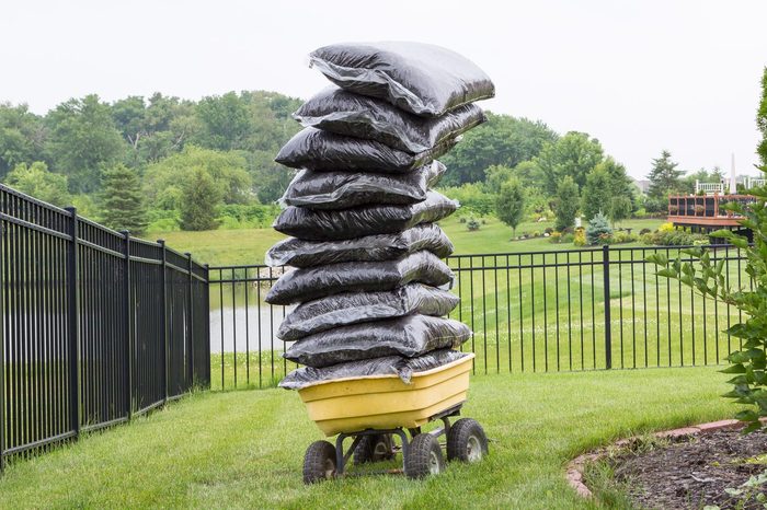 A yellow cart carries a tall stack of black bags, set against a green lawn, a fence, and a tranquil water feature in the background.