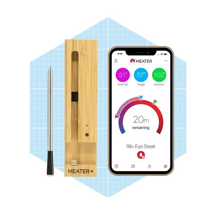 A digital meat thermometer with a bamboo holder and smartphone app displays cooking temperatures and time remaining for a steak.