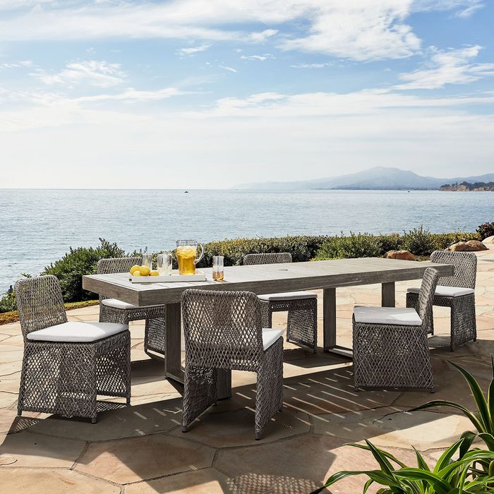 A rectangular dining table with woven chairs is set outdoors, accompanied by drinks and lemons, overlooking a calm sea and distant mountains under a clear sky.