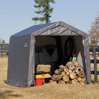 A grey storage shed stands in a grassy area, housing gardening tools and firewood, with a backdrop of trees and a fence under a clear sky.