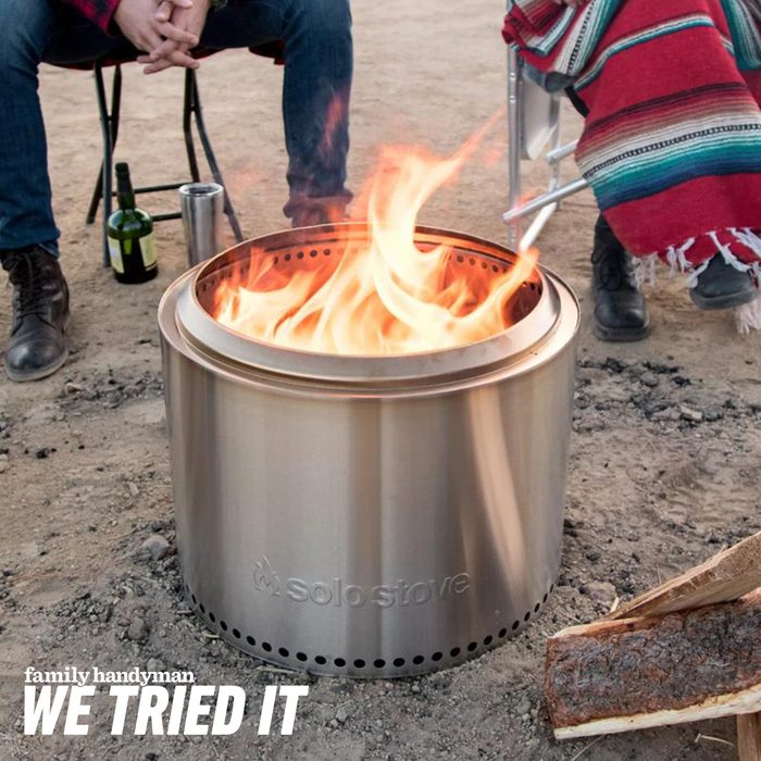 A metal fire pit burns brightly, surrounded by people sitting on chairs. Nearby, a bottle of wine and firewood are visible on the ground.