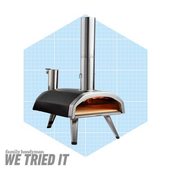 A portable pizza oven with a stainless steel chimney stands on sturdy legs, showcasing a warm, glowing interior. The background features a light blue grid pattern.