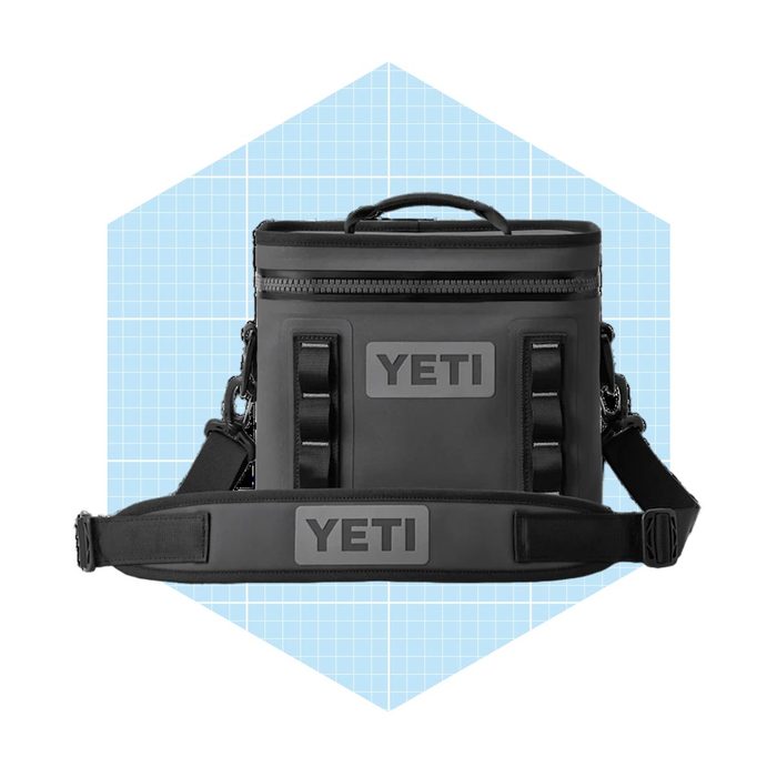 A black insulated cooler bag stands upright, featuring branded logos and a detachable shoulder strap against a light blue geometric background.