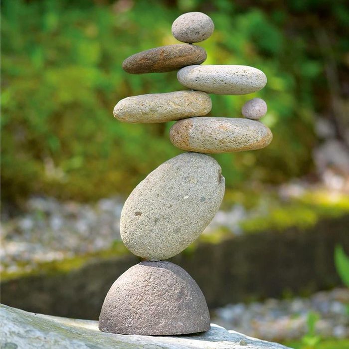 A stack of smooth stones balances on a larger stone, set against a blurred, natural green background with a soft stream visible nearby.