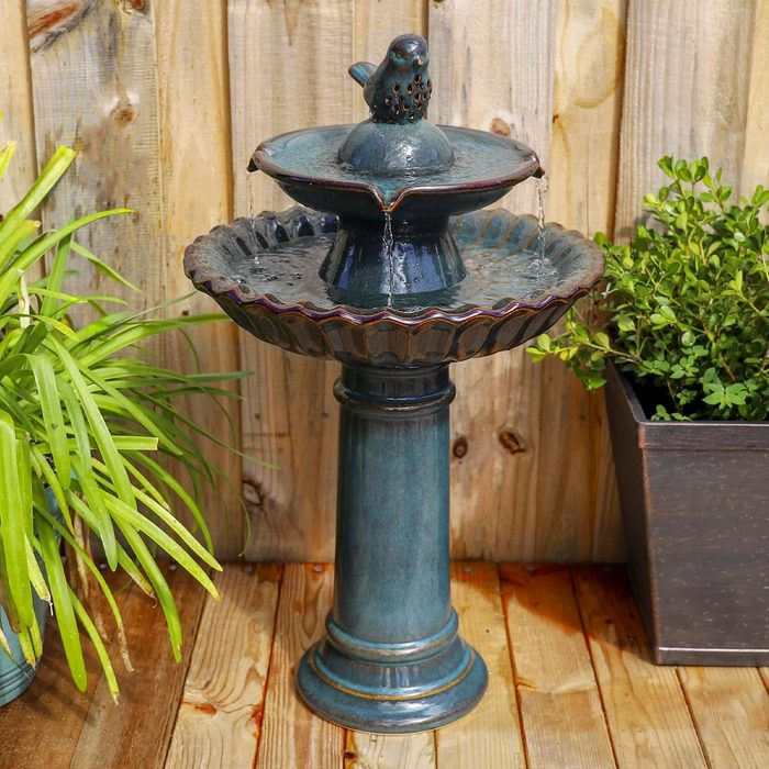 A decorative birdbath fountain stands on a wooden platform, water flows from a top basin to the bottom, surrounded by plants and a wooden backdrop.