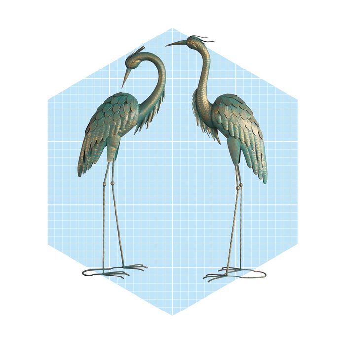 Two decorative metallic herons stand upright, one looking down and the other facing forward, set against a light blue grid-patterned background.