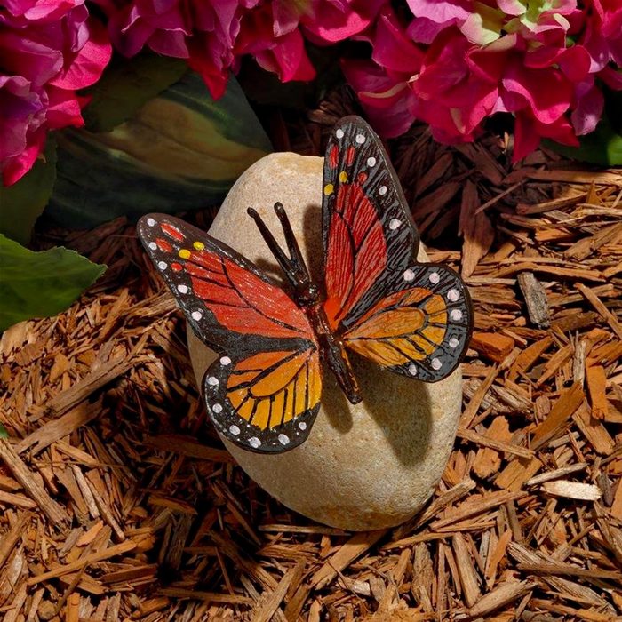A painted butterfly rests on a smooth stone, surrounded by mulch and vibrant pink flowers, creating a colorful and decorative garden scene.