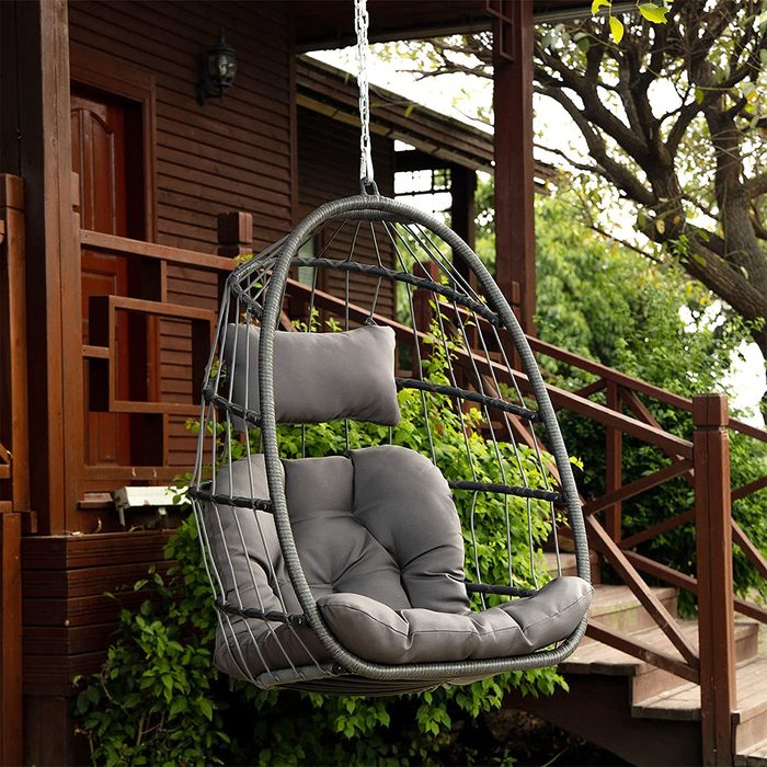 A hanging chair with gray cushions sways gently in a garden setting, surrounded by greenery and near a wooden staircase leading to a cabin.