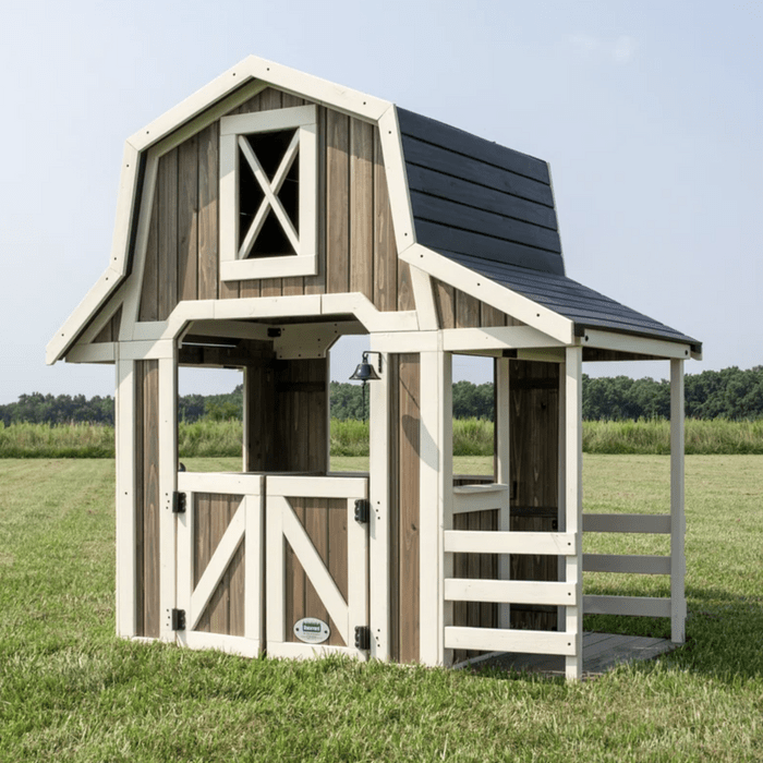A wooden playhouse resembling a barn stands on green grass. It features a gabled roof, open doors, and a bell, set against a scenic background.
