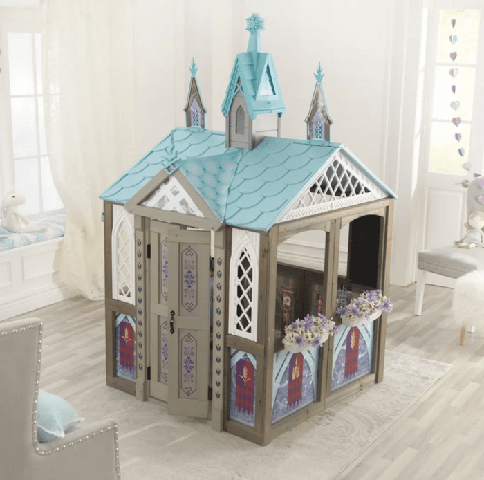 A colorful playhouse stands in a bright room, featuring a peaked blue roof, decorative windows, and flower boxes, inviting imaginative play.