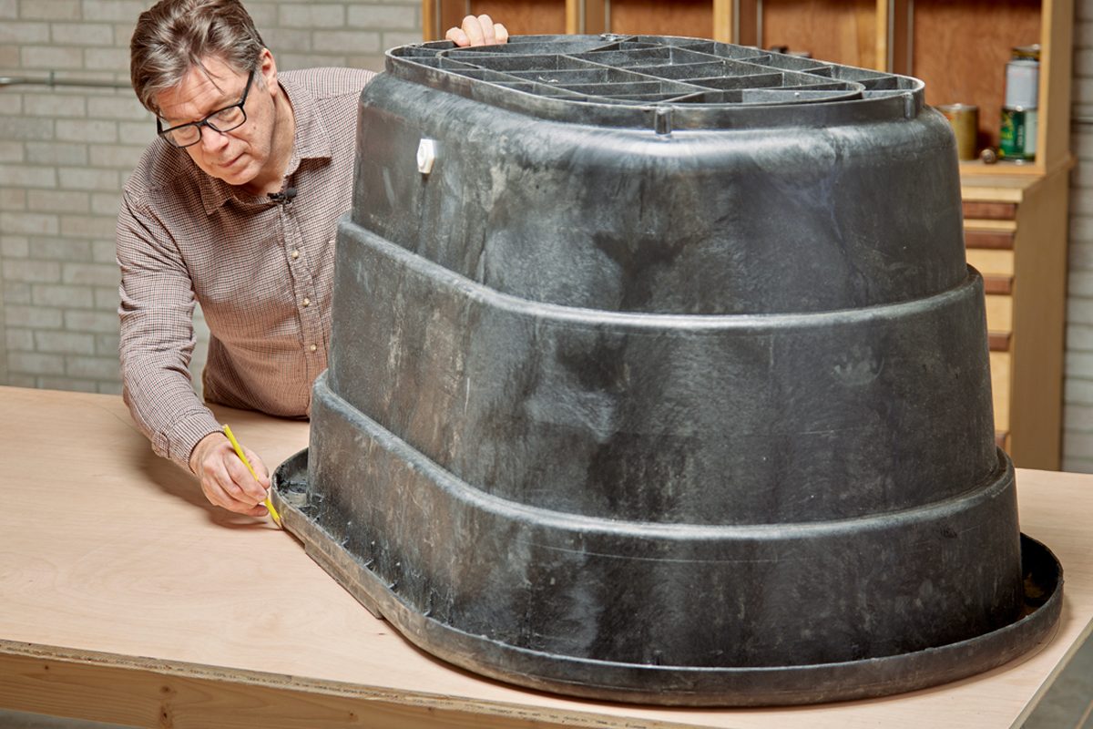 A man draws a line on a wooden surface while measuring the bottom of a large, dark plastic container in a workshop environment.