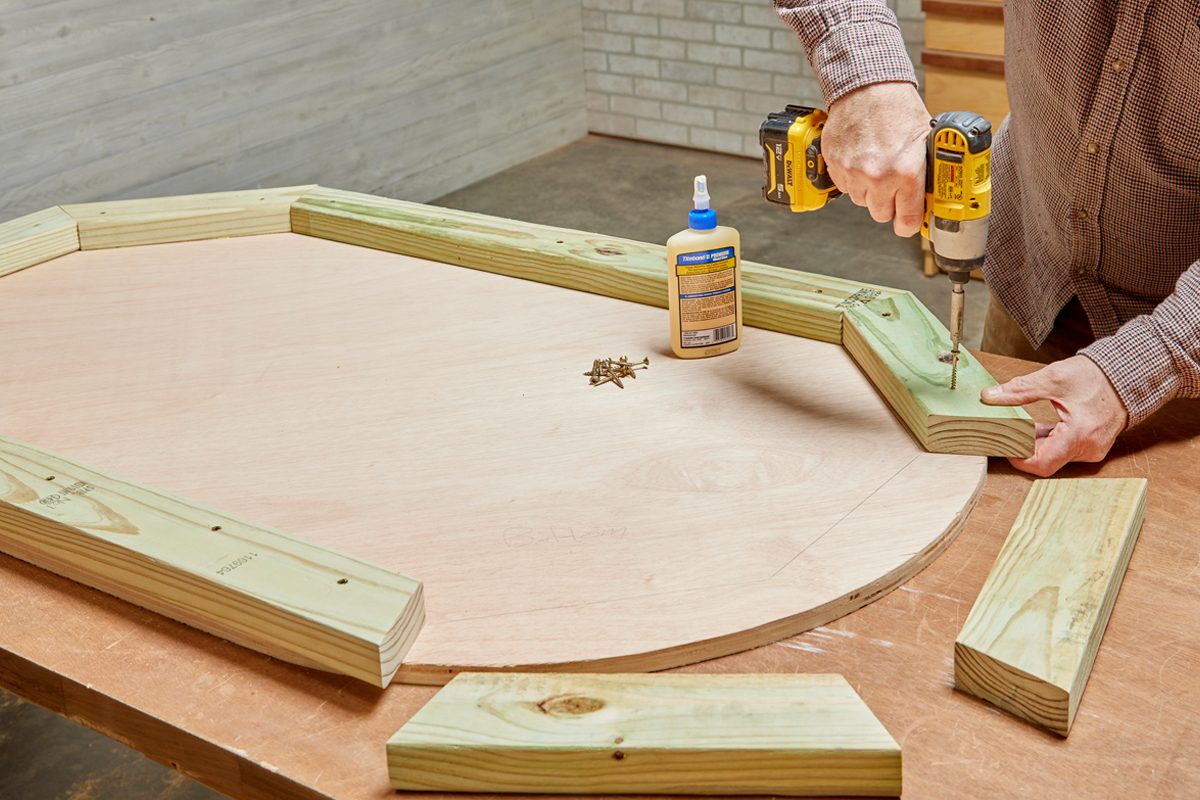 A person screws wooden pieces onto a circular plywood surface, using glue and a power drill, in a workshop with wooden walls and floors.