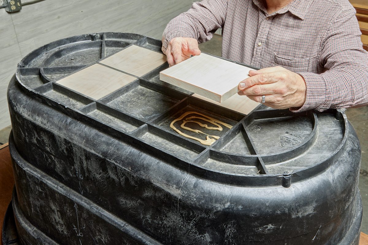 A person is placing a wooden panel onto a black, oval, plastic container, which has glue applied on its surface, in a workshop setting.