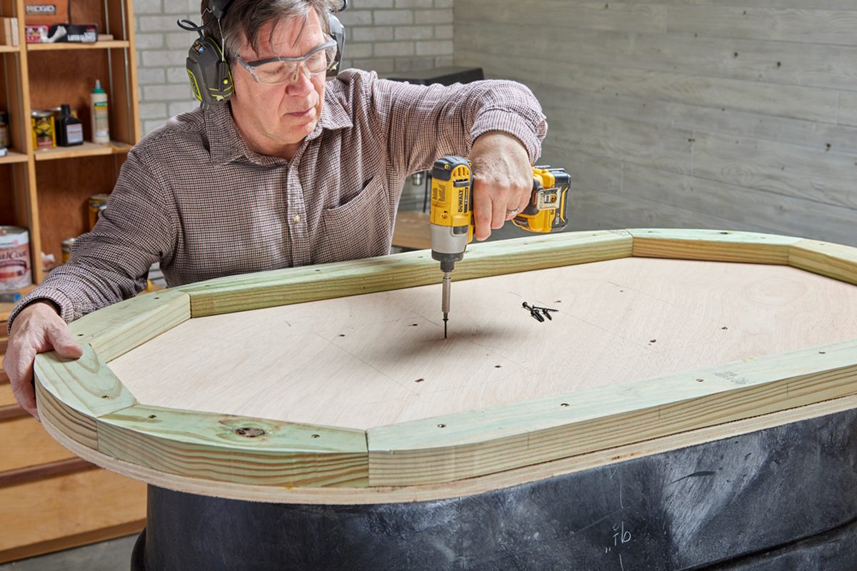 A man drills screws into an octagonal wooden piece, working at a black table in a well-lit workshop filled with tools and materials.