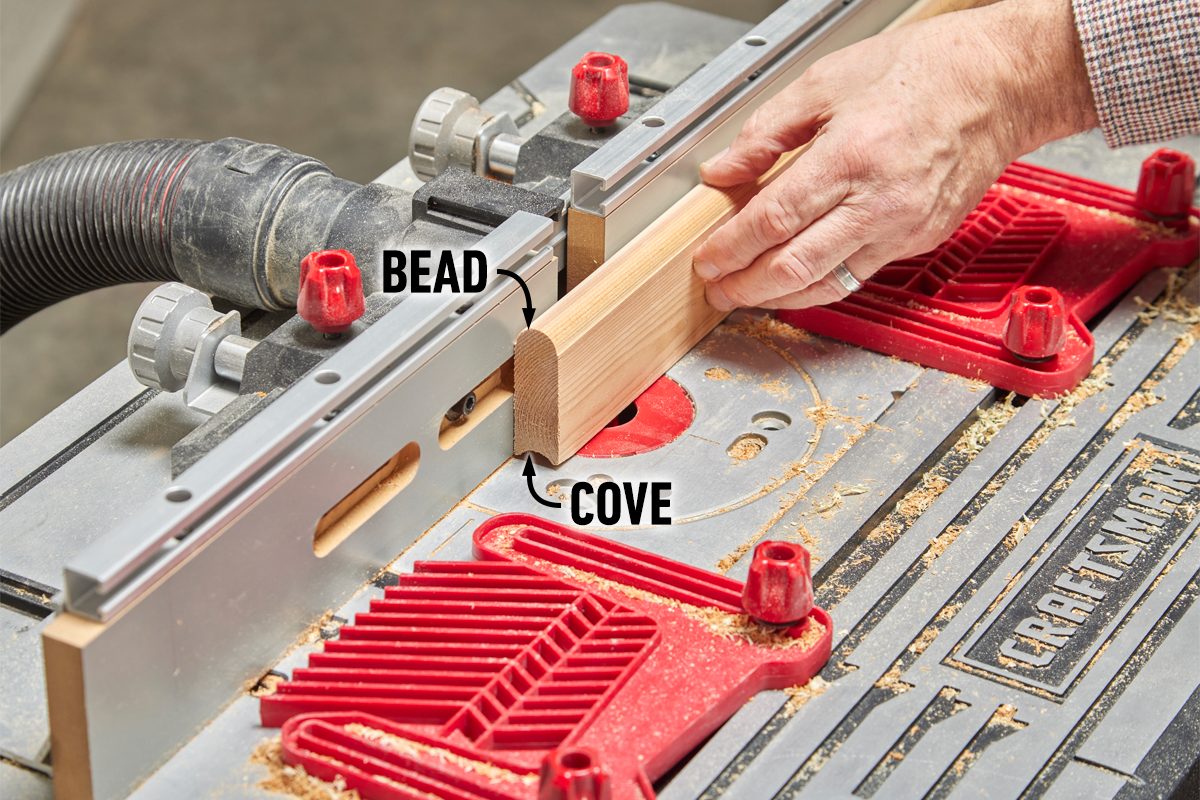 A hand guides a wooden piece through a router table, shaping it with a bead and cove pattern, surrounded by sawdust and tools.